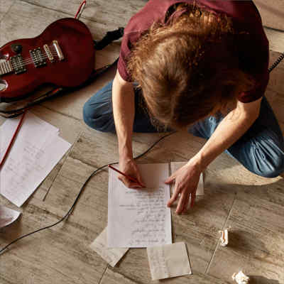 Songwriting lessons. A top down view of a person sitting on the floor handwriting on a sheet of paper.